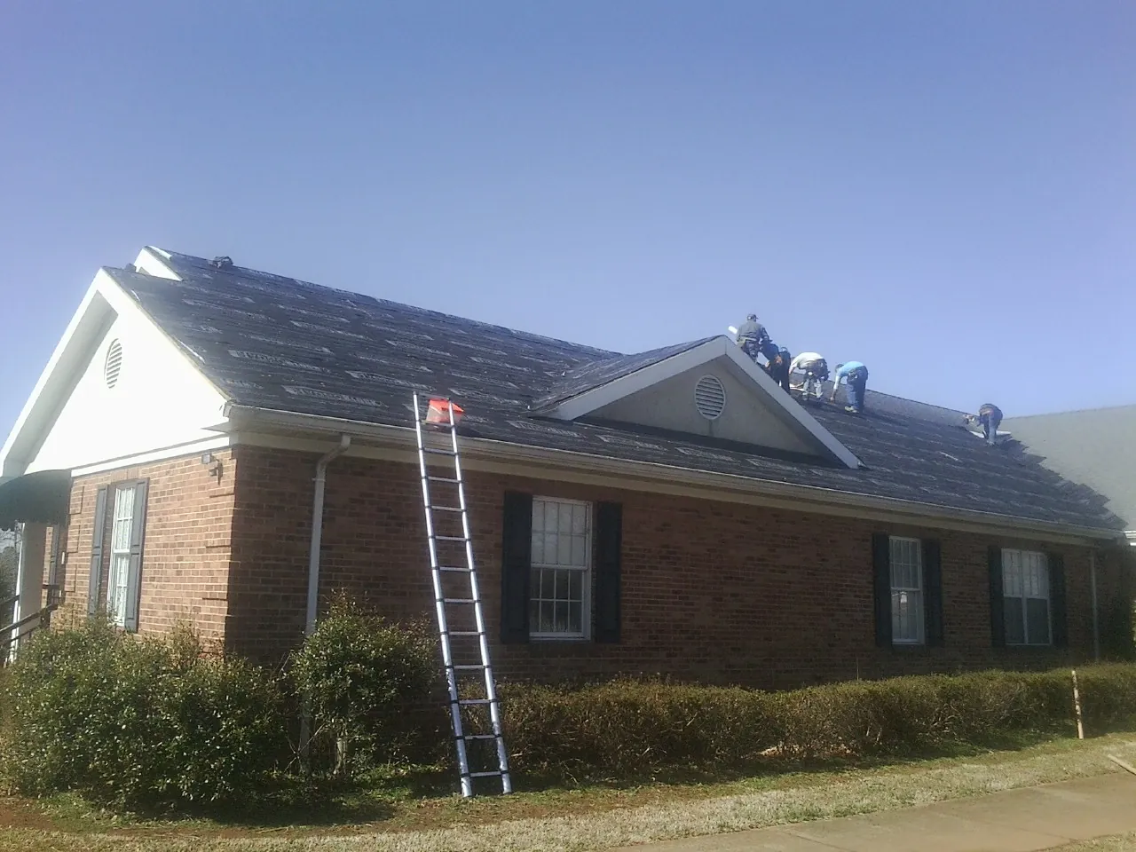 Crew tearing shingles off a brick ranch roof with a ladder set — Metal Roof Repair work by our crew in Pell Lake