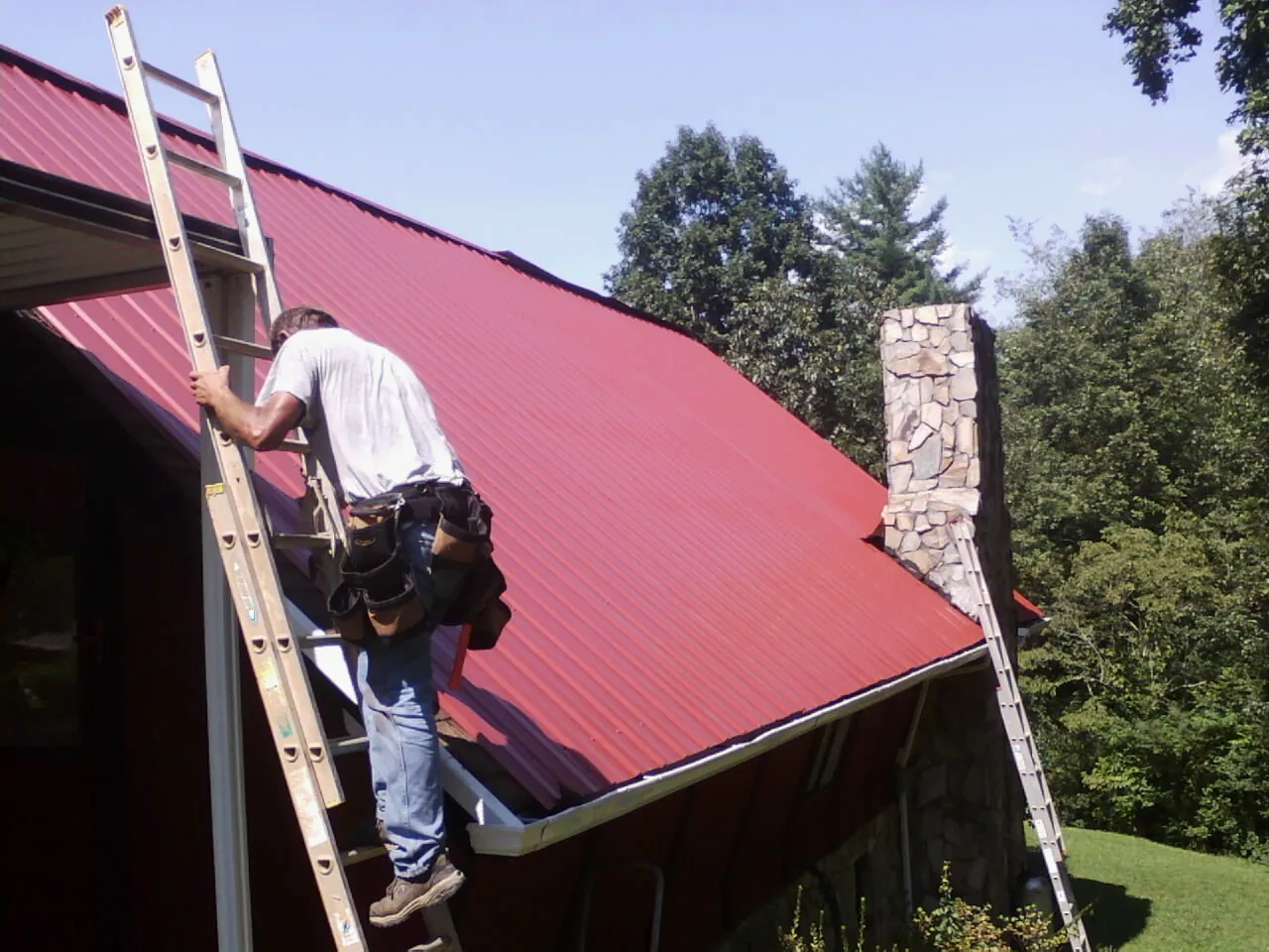 Roofer on a ladder beside a red metal roof and stone chimney — Soffit & Fascia Repair by Roofing Brothers in Pell Lake